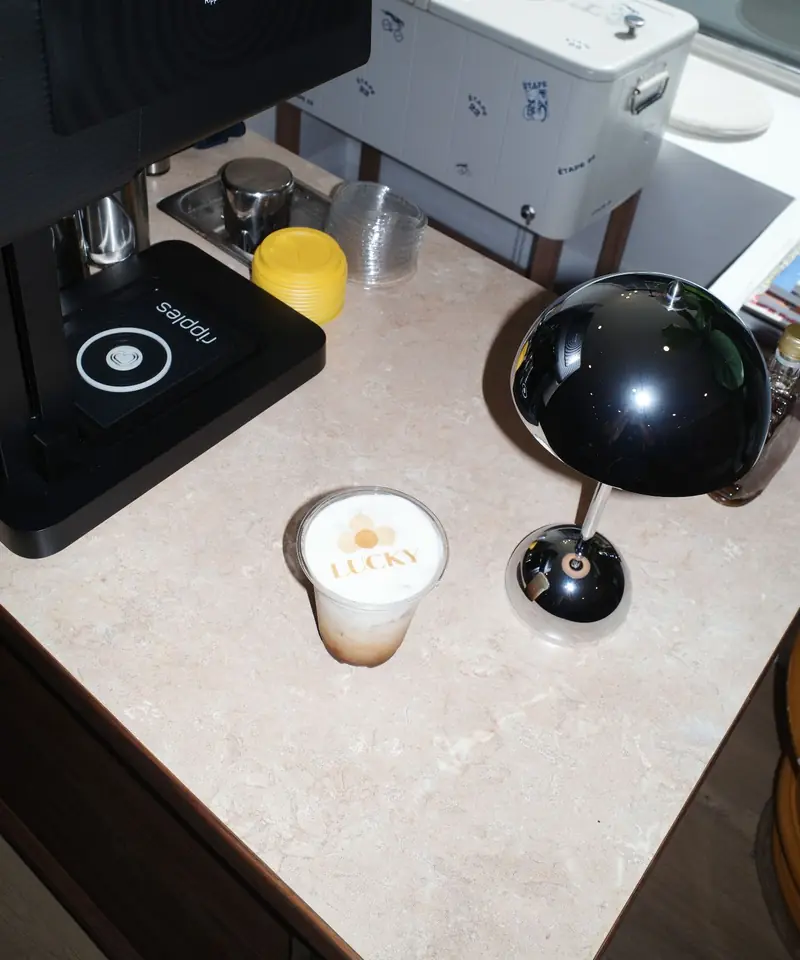 An iced latte with the Lucky logo printed on its foam beside the coffee printer and chrome lamp on the cart